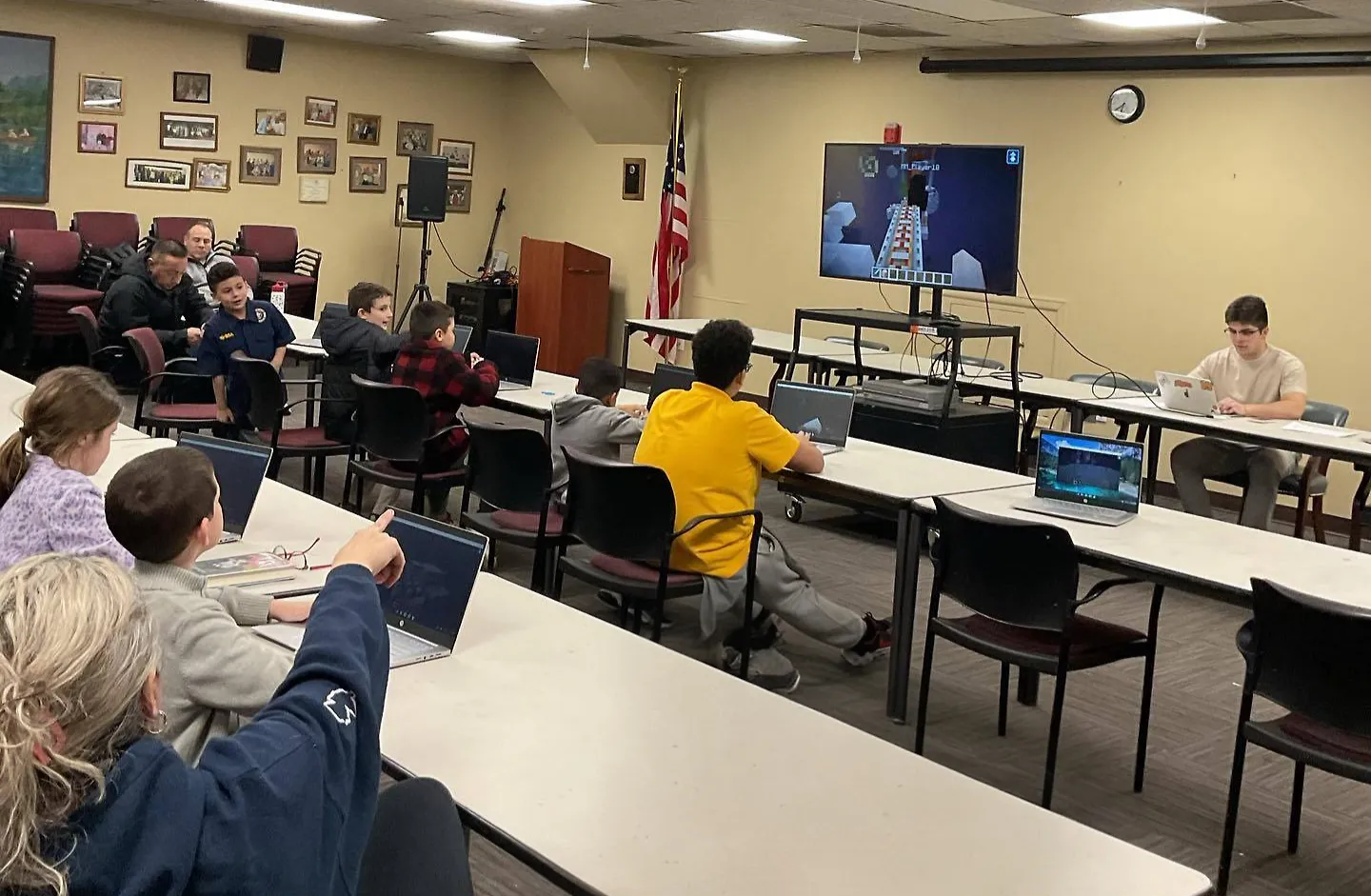 People sit in a classroom, using laptops, watching a gaming tutorial on a screen. A flag and framed photos decorate the beige walls.